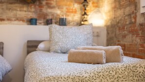 Een bed met een beddegoed met een patroon, twee opgerolde handdoeken, een bakstenen muur en een tafeltje met een lamp.