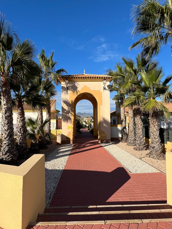 An arched entrance with a red brick pathway leading through it, surrounded by palm trees and buildings with yellow walls.