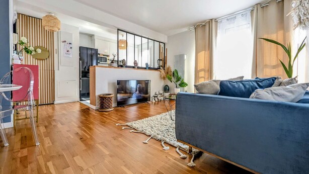 A modern living room with a blue sofa, wooden flooring, a TV, and a kitchen in the background.
