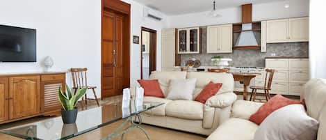 A modern living room with a glass coffee table, beige sofa, and a TV on the wall.