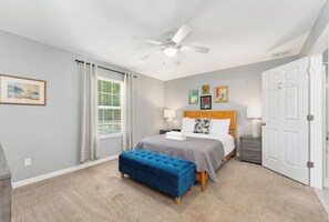 A bedroom with a bed, a ceiling fan, a window with blinds, a door, and a small table with a lamp.
