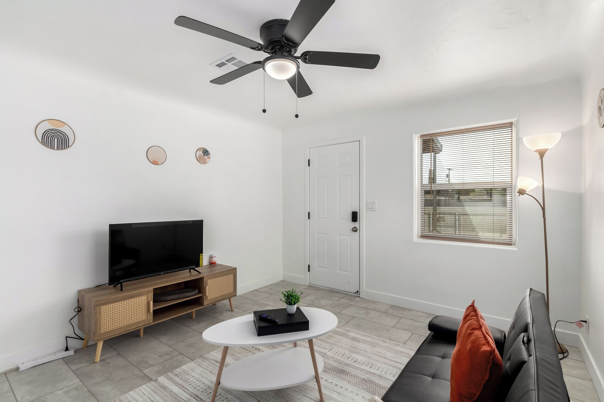 A modern living room with a flat-screen TV, a ceiling fan, a white door, and a window with blinds.