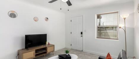 A modern living room with a flat-screen TV, a ceiling fan, a white door, and a window with blinds.