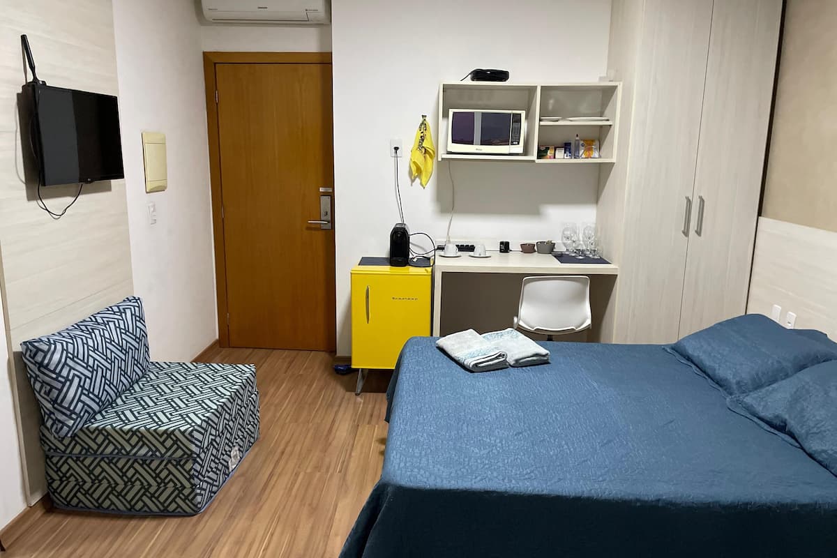 Quarto de hotel com cama, pequena mesa com micro-ondas, poltrona, sofá e guarda-roupa.