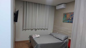 Un dormitorio pequeño y ordenado con una cama individual, una ventana con cortinas, un equipo de aire acondicionado y un tapiz colorido en la pared.