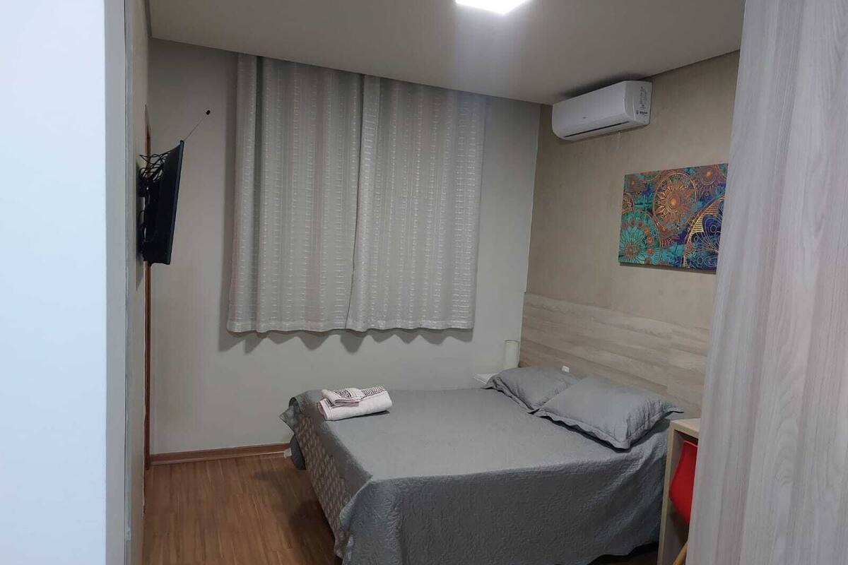 Quarto pequeno e bem organizado, com uma cama de solteiro, janela com cortinas, ar condicionado e uma tapeçaria colorida na parede.