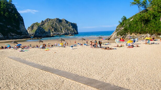 A beach with a wooden walkway, people relaxing, and a large rock formation in the background. - Tejasturias : La Franca Swimming Pool, Paddle Tennis and Beach (Noriega)