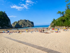 A beach with a wooden walkway, people relaxing, and a large rock formation in the background.