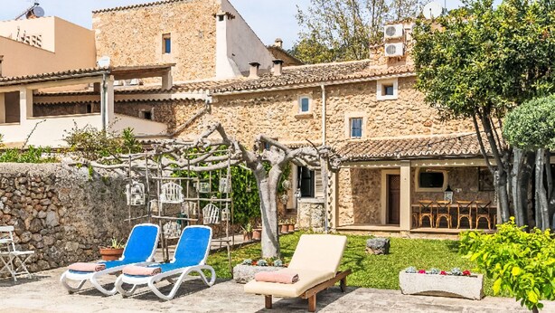 A poolside area with lounge chairs, a pergola with hanging birdhouses, and a stone building with a tiled roof.