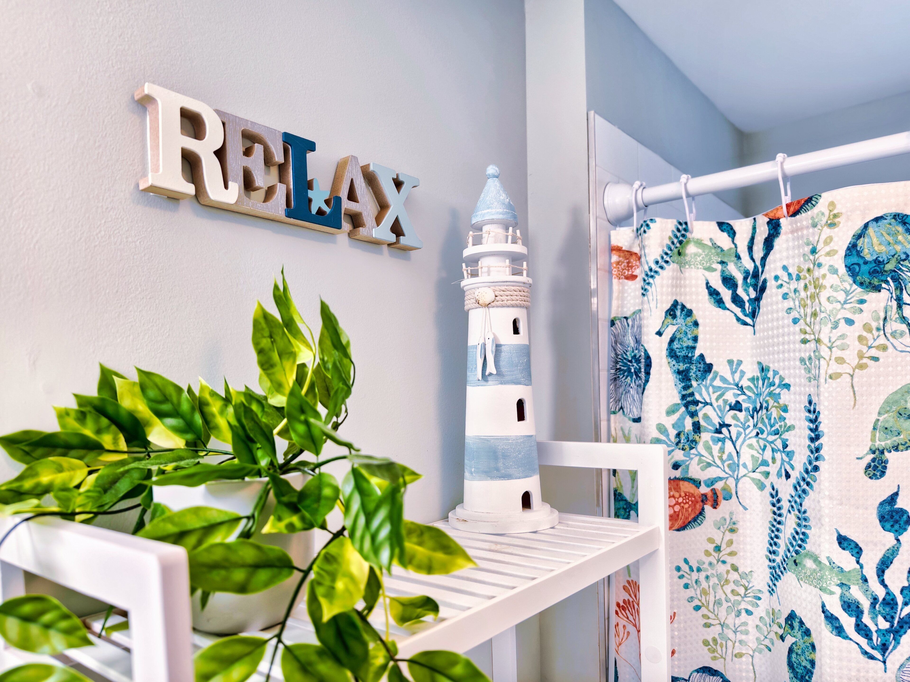 Une salle de bain avec une étagère blanche, une plante en pot, une décoration représentant un phare et un rideau de douche orné d’un motif aquatique bleu et vert.