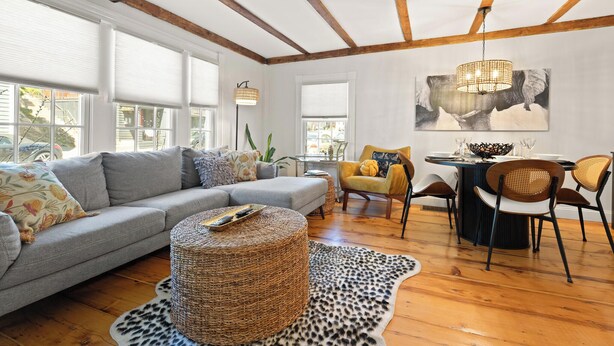 A cozy living room with a grey sofa, a round wicker ottoman, a leopard print rug, and a dining area with a table and chairs.