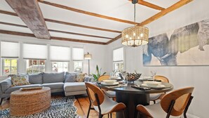 A modern living room with a grey sofa, wooden dining table, and chairs. There is a large window with blinds, a hanging light fixture, and a rug on the floor.