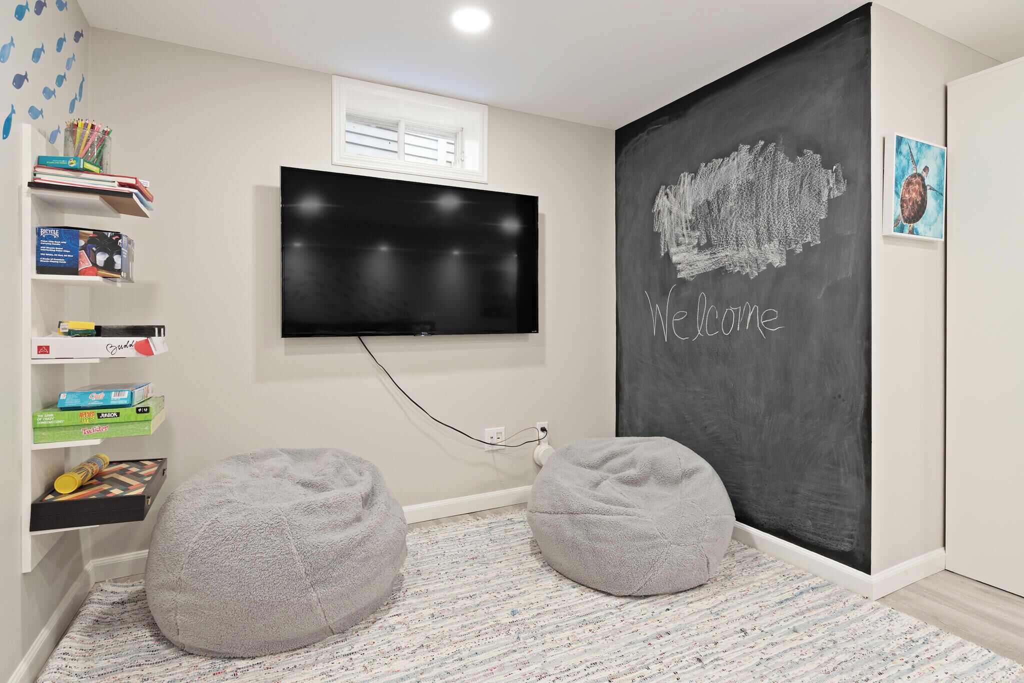 A room with a flat-screen TV mounted on the wall, a chalkboard with "Welcome" written on it, and two grey bean bags on a patterned rug.