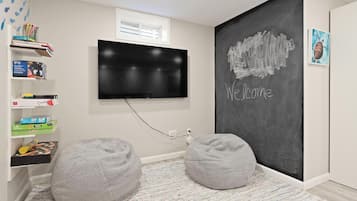 A room with a flat-screen TV mounted on the wall, a chalkboard with "Welcome" written on it, and two grey bean bags on a patterned rug.