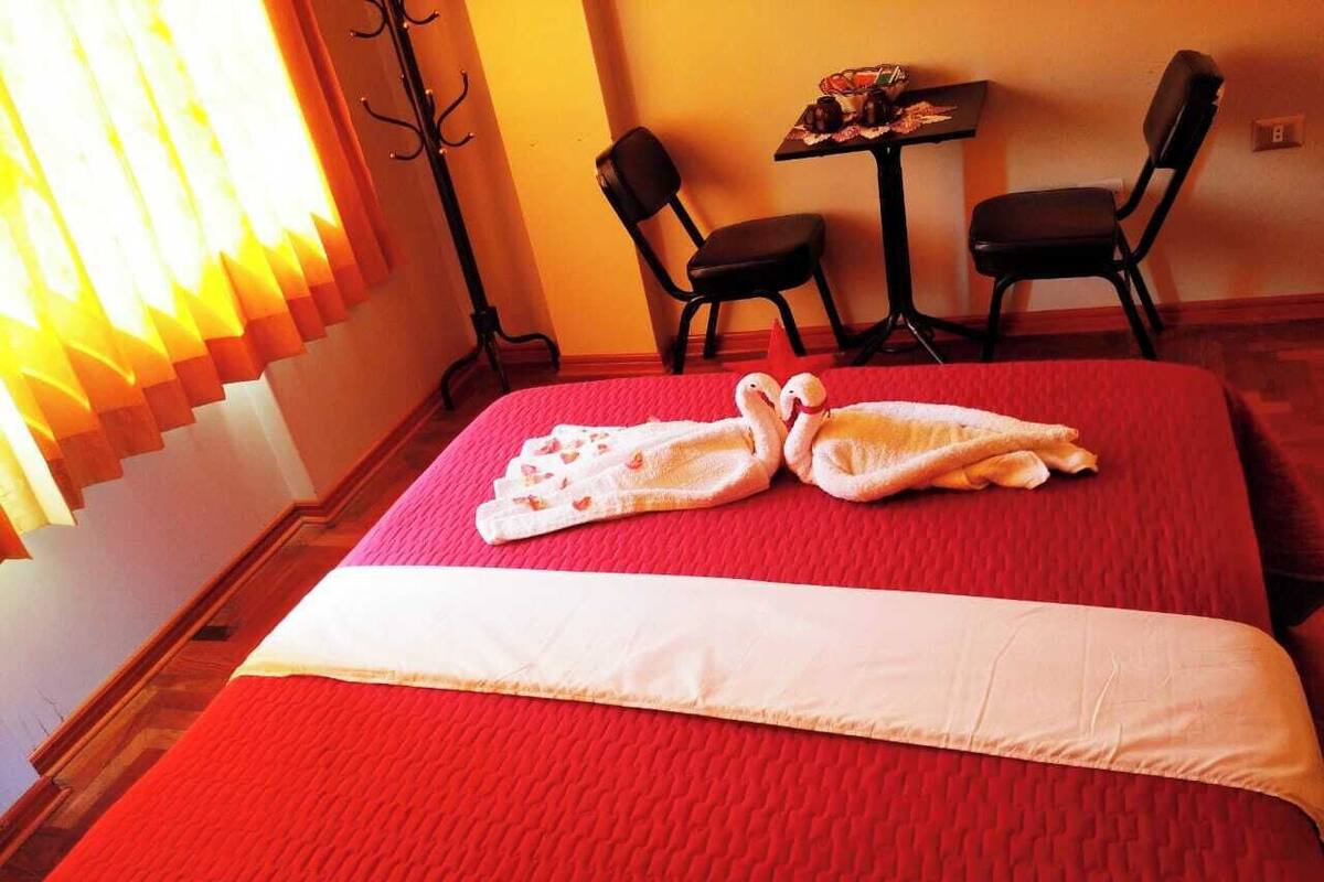 A hotel room with a red bedspread, two black chairs, a small table with a vase, and a window with curtains.