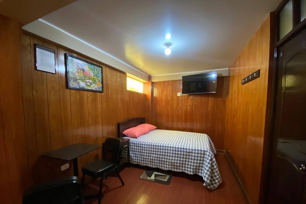 A small, cozy room with wooden walls, a single bed with a checkered bedspread, a desk with a chair, and a television mounted on the wall.