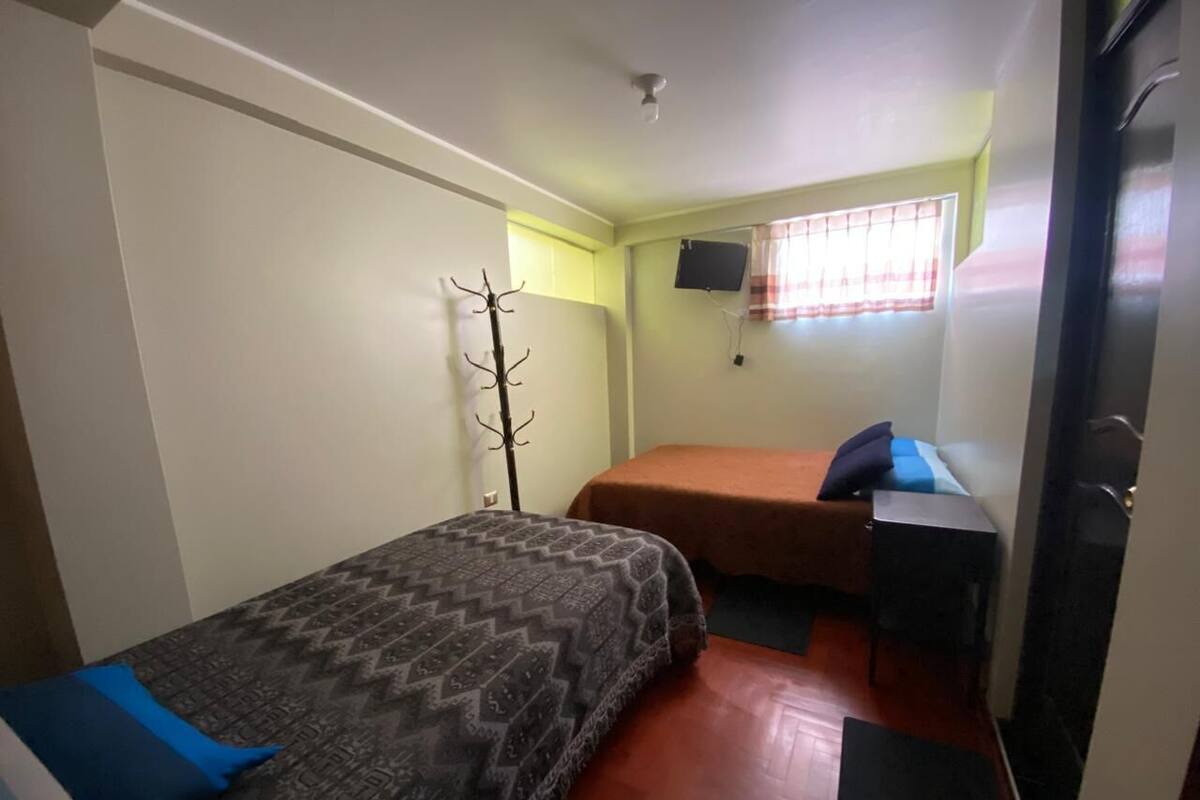 A small, compact room with two beds, a single chair, a coat rack, and a window with a curtain.