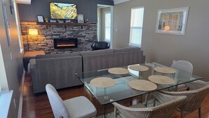 A modern living room with a stone fireplace, a large flat-screen TV, a glass dining table with woven placemats, and wicker chairs. - Ocean Pearl-Beach block, Quick stroll to Boardwalk (Atlantic City)