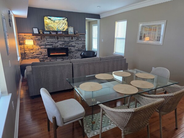 A modern living room with a stone fireplace, a large flat-screen TV, a glass dining table with woven placemats, and wicker chairs.