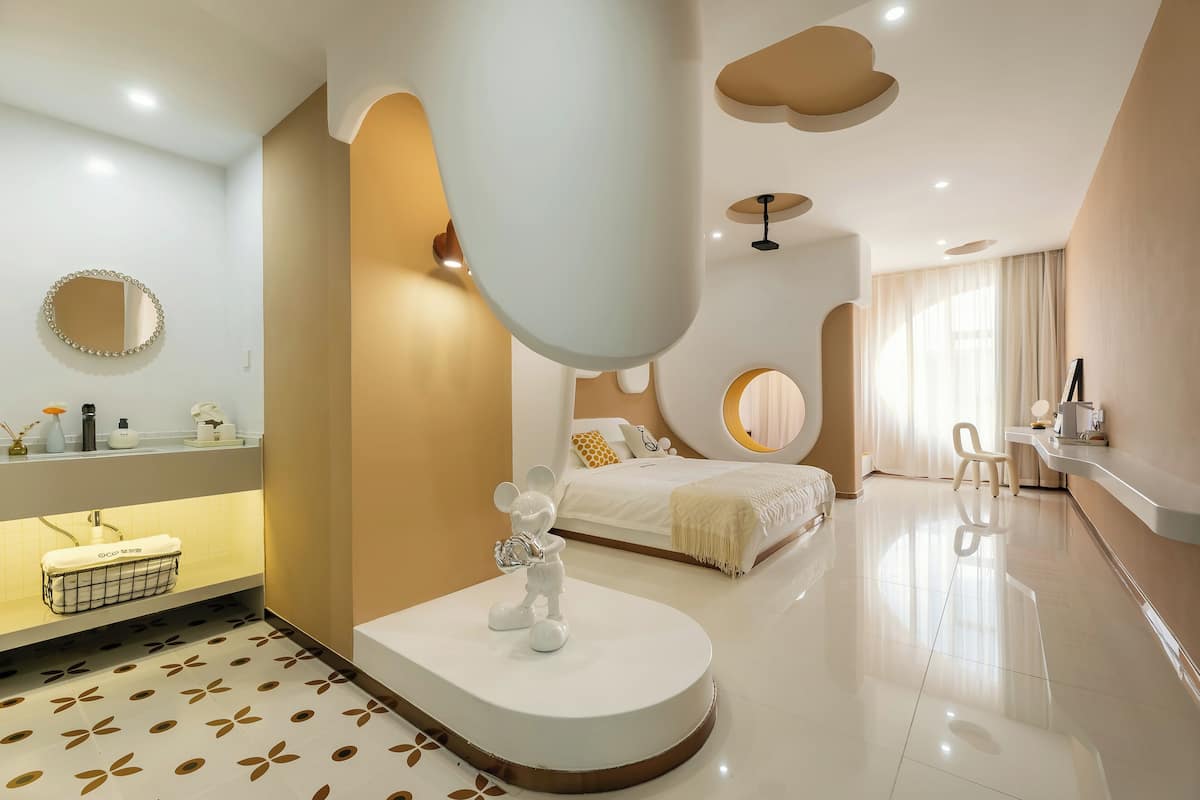 Um quarto moderno com uma estátua do Mickey Mouse, um espelho redondo, uma cama com cabeceira branca e um banheiro com pia e espelho brancos.