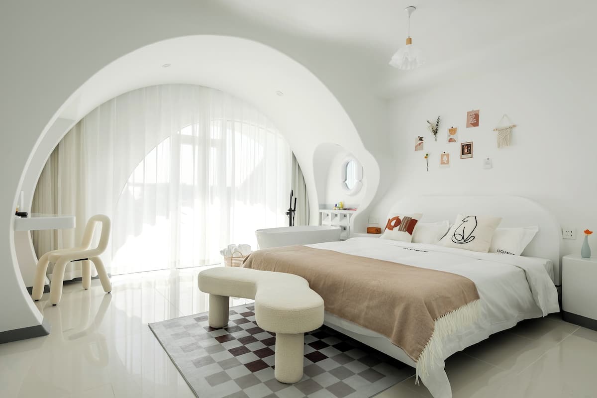 Quarto moderno com uma grande janela em arco, cama com manta bege, escrivaninha branca e uma cadeira.