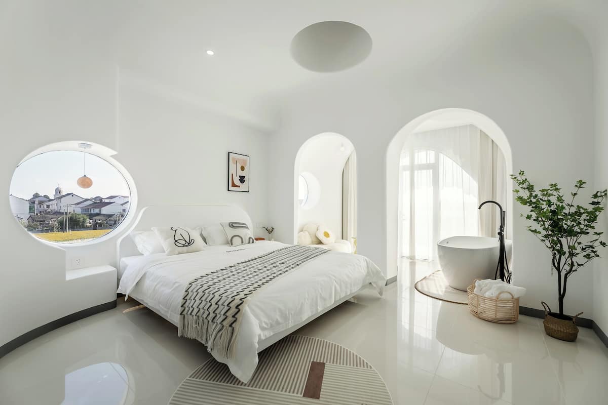 Quarto moderno com uma cama grande, uma janela redonda, uma banheira e um vaso de planta.
