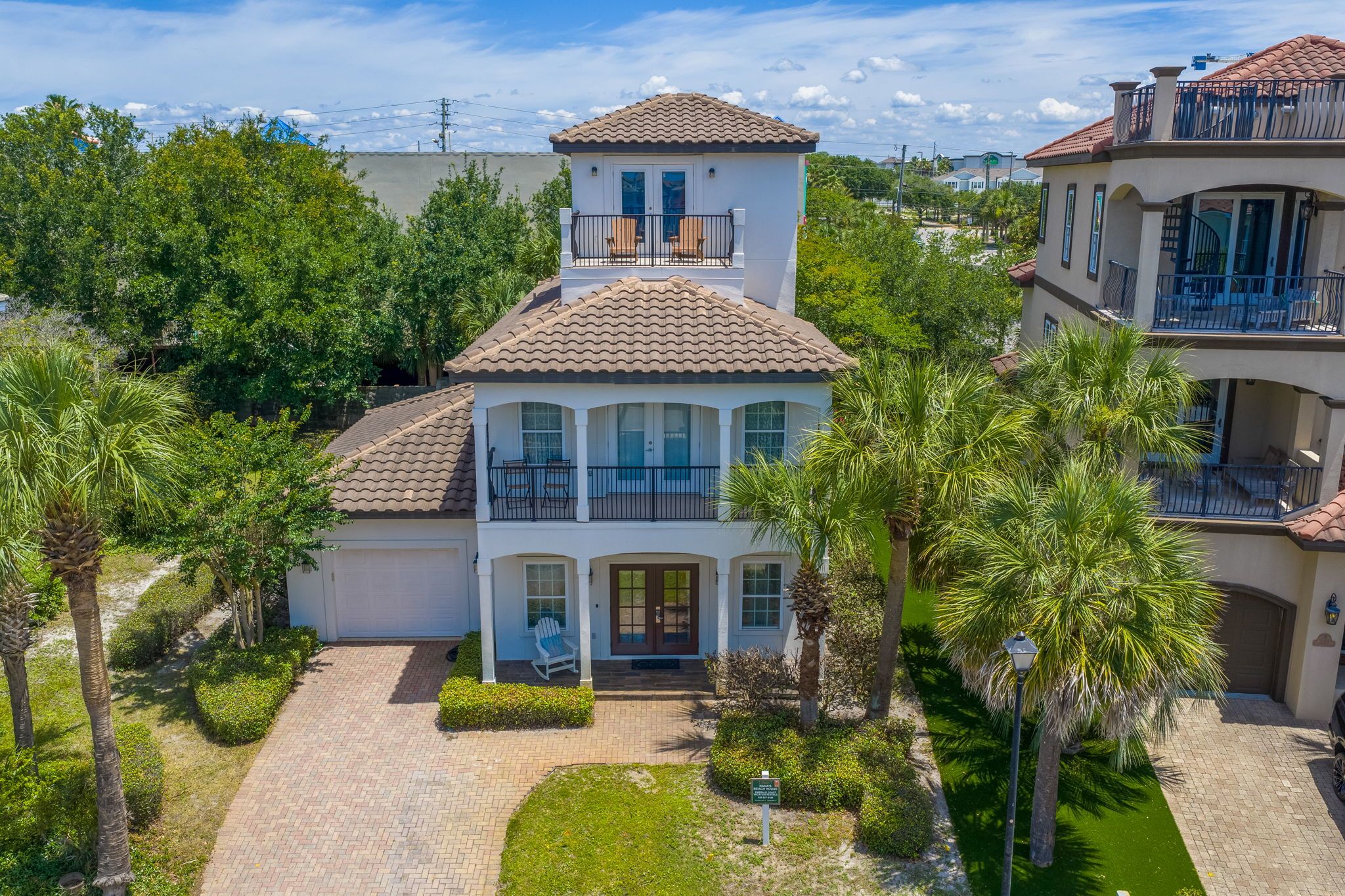 A two-story house with a balcony, surrounded by palm trees and a paved driveway.