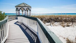 Ein hölzerner Steg führt zu einem weißen Gazebo mit Bögen, das auf einen Strand mit Sand und spärlicher Vegetation blickt.