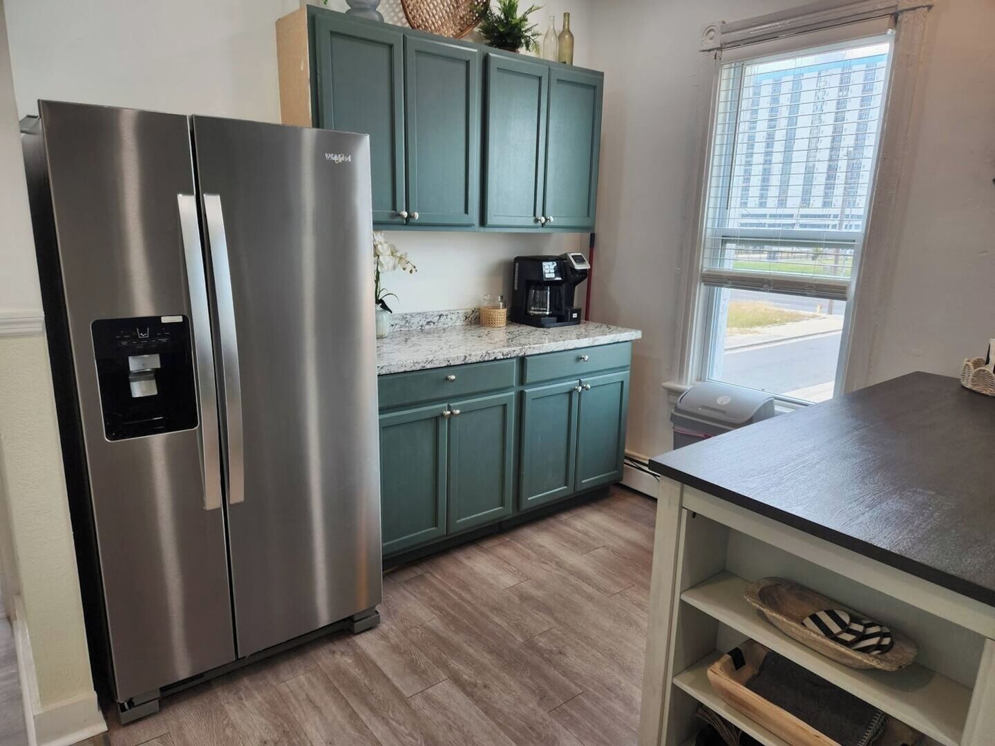 A kitchen with teal cabinets, a stainless steel refrigerator, a coffee maker, and a window with a view of buildings.