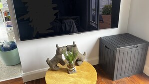 A room with a large mirror, a small round table with a plant, a grey trash bin, and a wooden floor.