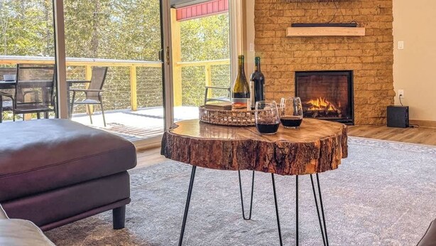 A modern living room with a stone fireplace, a round wooden table with wine glasses and bottles, and a large TV mounted on the wall. - Family Retreat | Game Room + EV Charger (Sturgeon Bay)