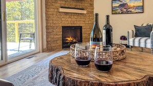 A cozy living room with a fireplace, a television mounted on a brick wall, and a dining table with wine glasses and a bottle. - Family Retreat | Game Room + EV Charger (Sturgeon Bay)
