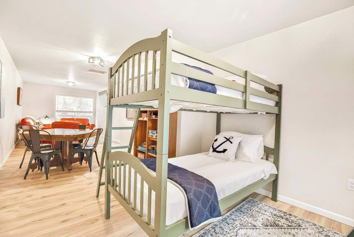 A bunk bed with a lower bunk, a desk with chairs, a dining table with chairs, and a patterned rug.
