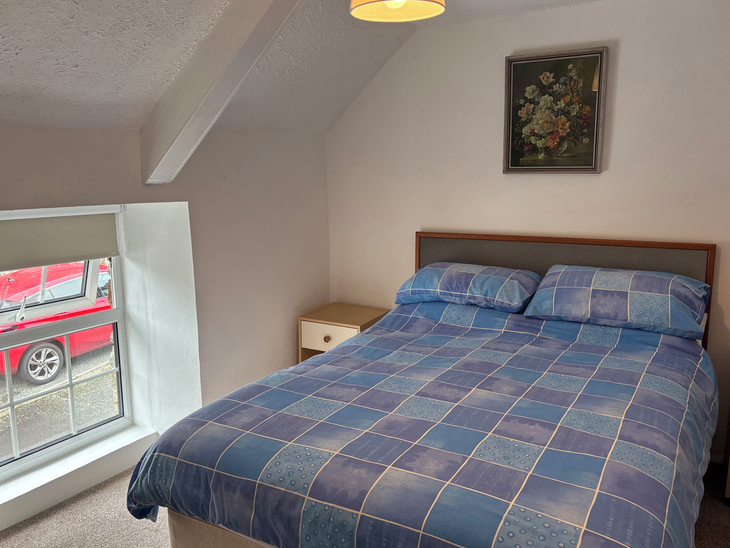A bedroom with a bed, a nightstand, a window with a view of a red car, and a framed picture on the wall.
