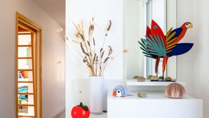 Un intérieur moderne avec une console blanche, un vase contenant des plantes séchées, une décoration représentant un perroquet aux couleurs vives et une sculpture en forme de tomate rouge.
