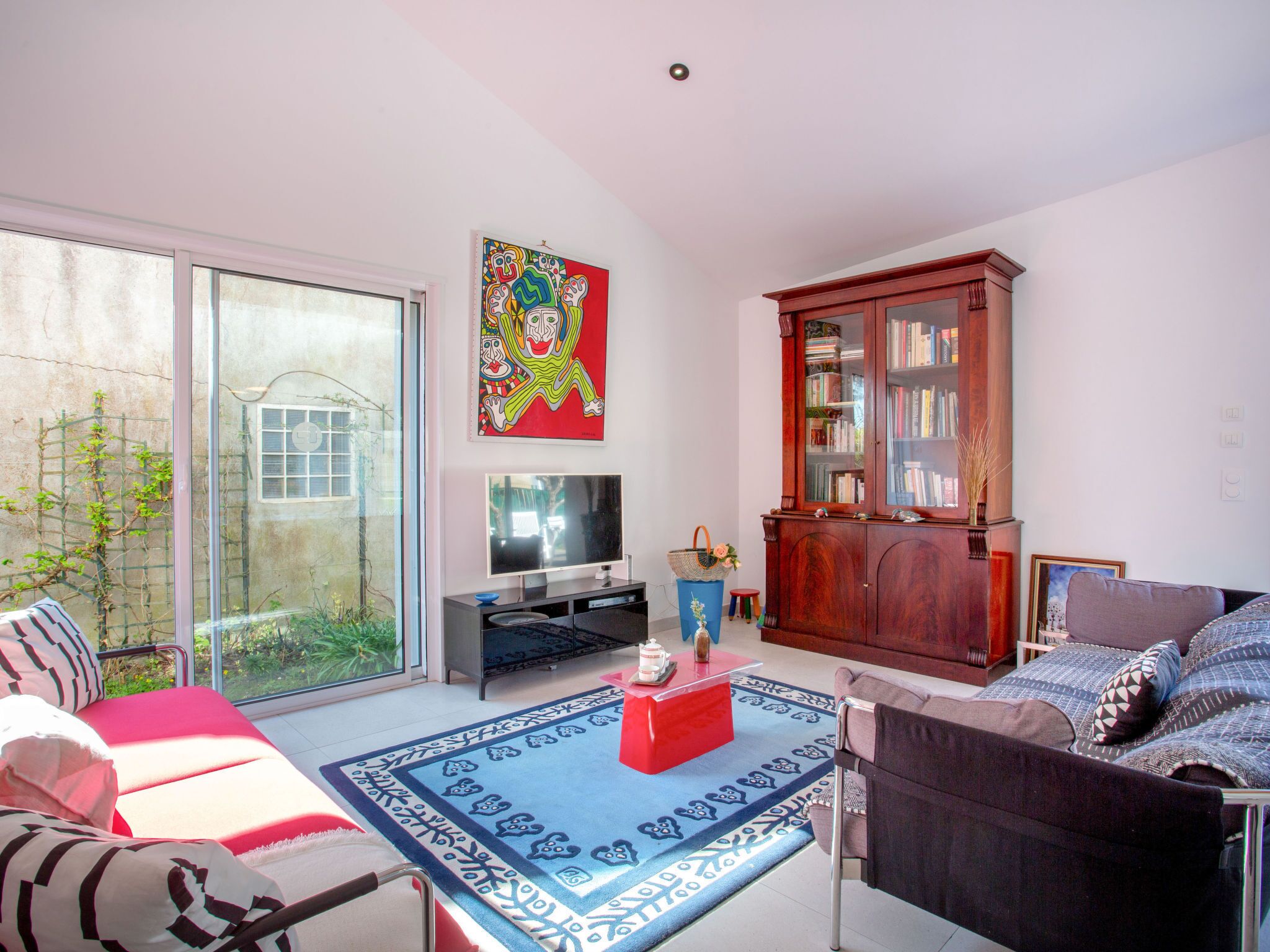 Een woonkamer met een bank, een rode salontafel, een televisie, een boekenkast en een groot raam.