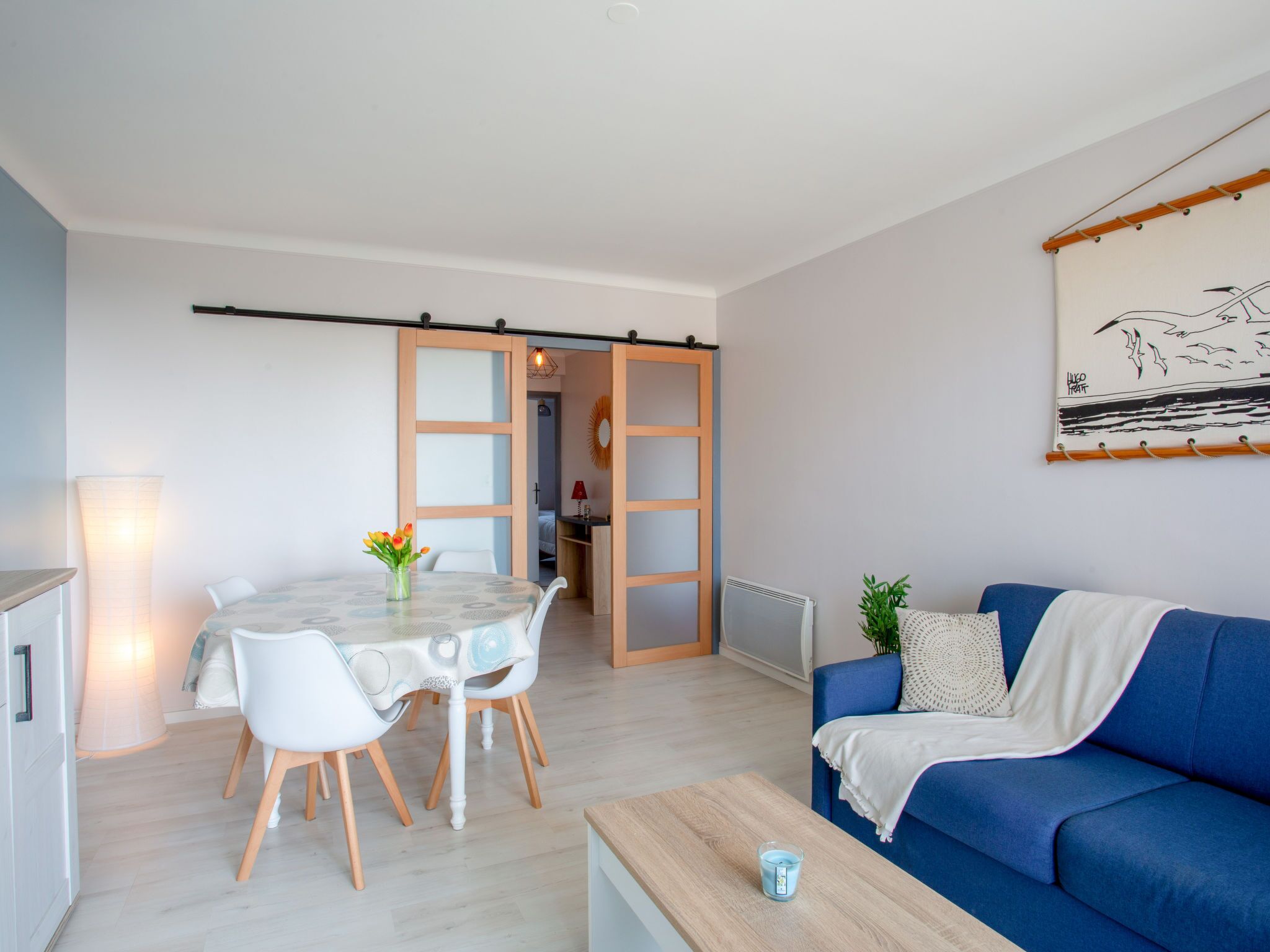 A modern living room with a blue sofa, a dining table with chairs, and a kitchen area visible through an open door.