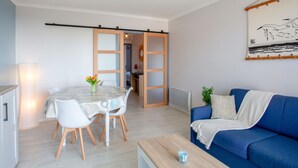 A modern living room with a blue sofa, a dining table with chairs, and a kitchen area visible through an open door.