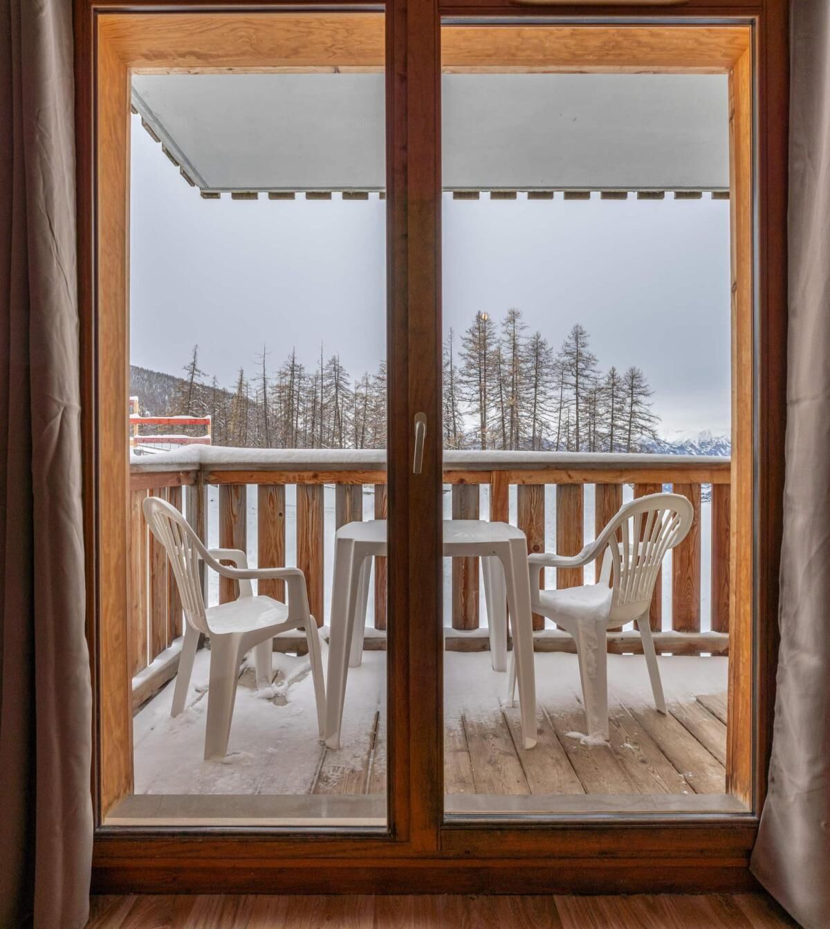 Een houten terras met een glazen deur die uitkomt op een besneeuwde omgeving, met stoelen en uitzicht op bomen en bergen.