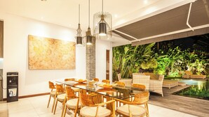 Un comedor moderno con una mesa de vidrio y sillas de madera, un cuadro abstracto grande y vistas a un exuberante jardín interior y piscina.