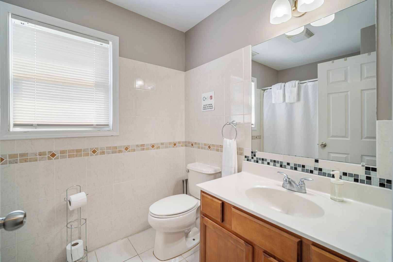 Een badkamer met toilet, wastafel en spiegel. Er is een raam met zonwering, een handdoekhouder en een bord op de muur.
