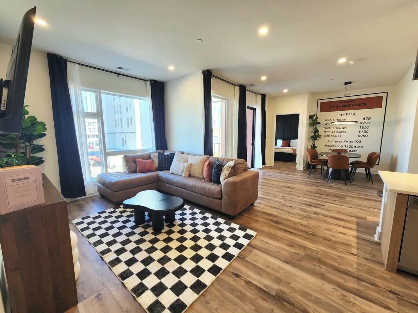 Un salon moderne avec un canapé d’angle, une table basse et un tapis à carreaux. On y trouve également un coin repas avec des chaises et une table, ainsi qu’un panneau d’affichage contenant des informations sur les locations.