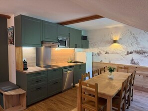 Eine kleine Küche mit modernen Geräten, ein Essbereich mit Holzmöbeln und ein Wandbild mit Bergmotiv.