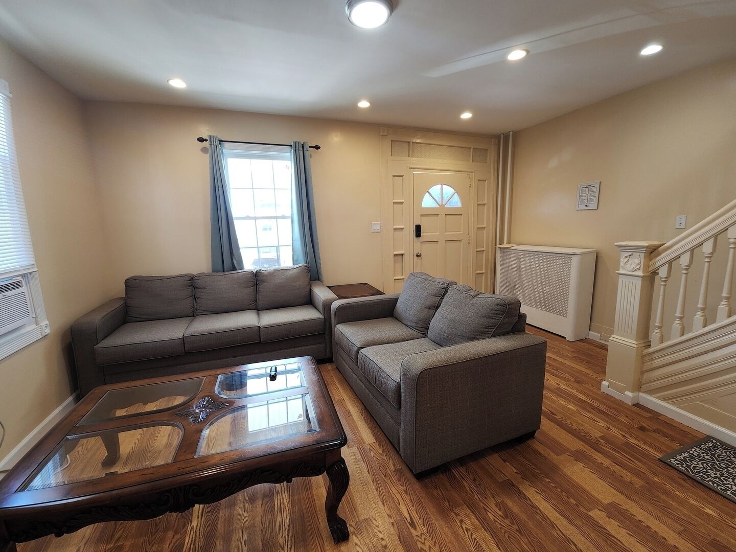 A living room with two grey sofas, a glass-top coffee table, wooden flooring, a radiator, and a staircase.