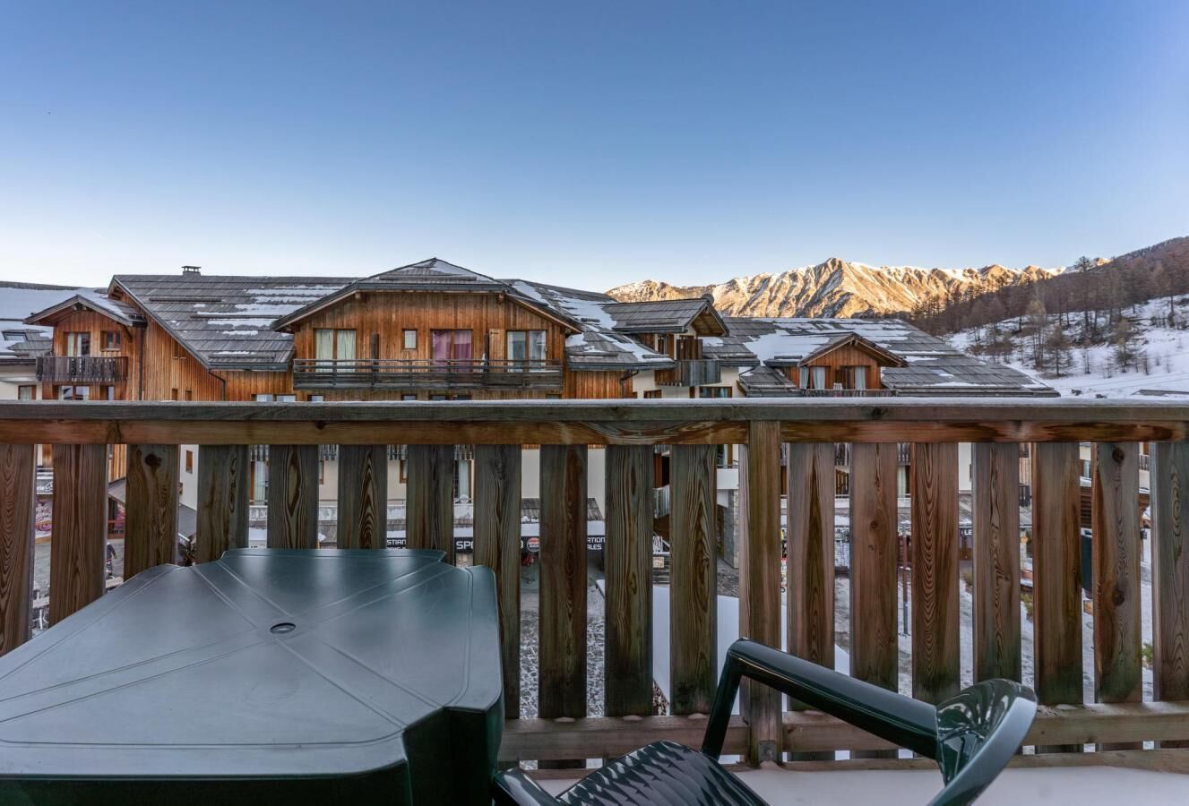 A wooden deck with a table and chairs, overlooking a row of chalet-style buildings with snow on the roofs, set against a mountainous backdrop.