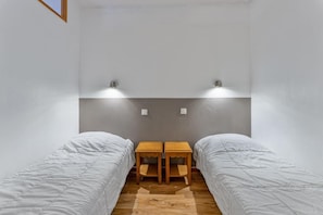 A small, modern bedroom with two single beds, a wooden nightstand, and a wall-mounted light fixture.
