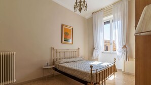 Une chambre à coucher avec un lit, un radiateur, une petite table, un lustre et une fenêtre avec des rideaux.