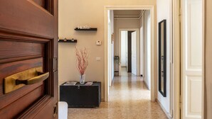 Un couloir avec une porte en bois, une valise noire, un vase avec des branches, une étagère sur laquelle est posé un panneau indiquant « MAISON », et un miroir au mur.