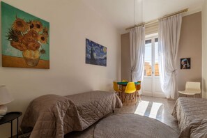 Une chambre avec deux lits, une table à manger avec des chaises, un tableau représentant des tournesols et une fenêtre avec des rideaux.