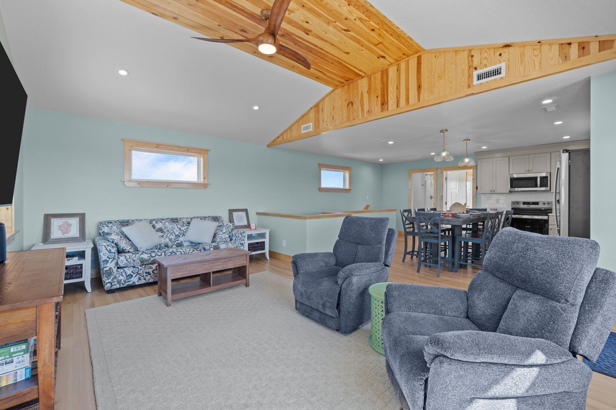 A spacious living area with a high ceiling, wooden beams, a sofa, two armchairs, a coffee table, and a dining area with a kitchen in the background.